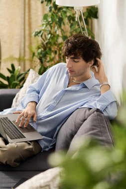 Kıvırcık saçlı yakışıklı genç adam canlı ve modern bir dairede dizüstü bilgisayarına odaklanıyor..