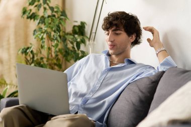 Kıvırcık saçlı yakışıklı genç adam kanepede rahatlıyor, bilgisayarını içeride meşgul ediyor..