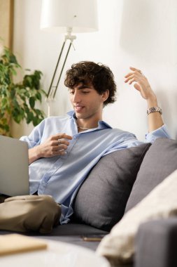 Kıvırcık saçlı genç bir adam şık dairesinde bir kanepede dinleniyor, dizüstü bilgisayarına odaklanıyor..