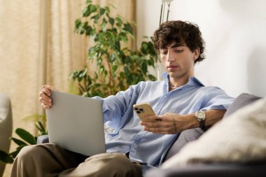 Yakışıklı genç adam rahat bir kanepede rahatlarken telefonunu ve laptopunu kullanıyor..