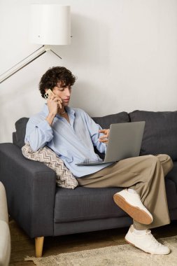 Mavi tişörtlü, kıvırcık saçlı genç adam dizüstü bilgisayarı üzerinde çalışırken telefonda konuşuyor..