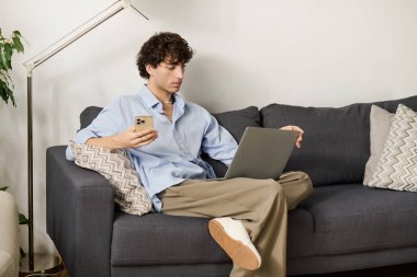 Kıvırcık saçlı genç bir adam modern bir dairede dizüstü bilgisayarını ve telefonunu kullanarak rahatlıyor..