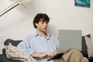 Kıvırcık saçlı şık bir genç adam rahat bir dairede oturur ve dizüstü bilgisayarına odaklanır..