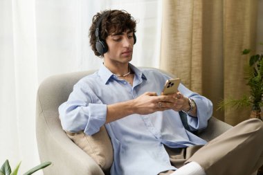 Kıvırcık saçlı genç adam pelüş bir sandalyede dinleniyor, telefonuna odaklanıyor ve anın tadını çıkarıyor..