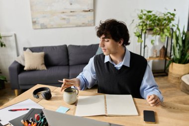 Kıvırcık saçlı genç bir adam masada oturur, defterine odaklanırken kahvesini yudumlar..
