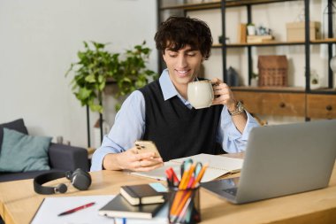 Kıvırcık saçlı yakışıklı genç adam dairesinde telefonunu kullanırken kahveyle rahatlıyor..
