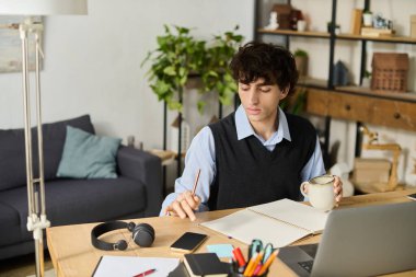 Kıvırcık saçlı genç bir adam masaya oturur, elinde kupa tutarken işine odaklanır..