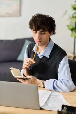 Kıvırcık saçlı yakışıklı genç bir adam modern dairesinde oturuyor ve defterini inceliyor..