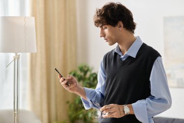 Kıvırcık saçlı yakışıklı genç bir adam şık dairesinde dikilirken telefonuna bakar..