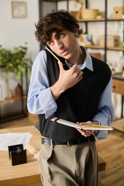 Mavi gömlekli yakışıklı genç bir adam elinde bir defter tutarken telefonda..