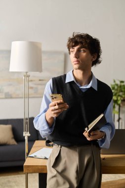 Yakışıklı bir genç adam, elinde telefon ve defterle modern dairesinde düşünceli bir şekilde duruyor..