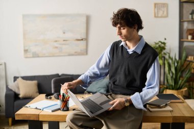 Mavi tişörtlü genç adam rahat bir ev ortamı ile çevrili olan dizüstü bilgisayarına odaklanıyor..