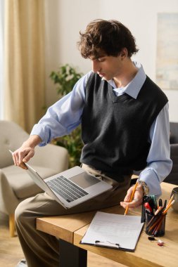 Kıvırcık saçlı genç adam rahat bir dairedeki masada otururken dizüstü bilgisayarına odaklanıyor..