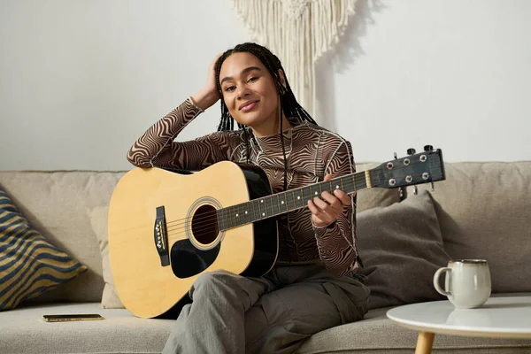 Saçları örgülü genç bir Afrikalı Amerikalı kadın rahat dairesinde gitar çalıyor, neşe saçıyor..