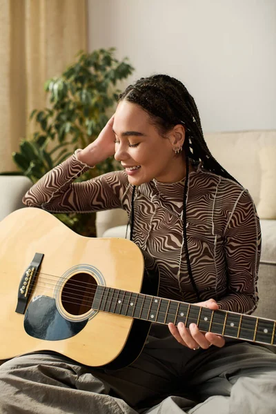 Saçları örgülü genç bir Afrikalı Amerikalı kadın rahat dairesinde otururken gitar çalmaktan hoşlanıyor..