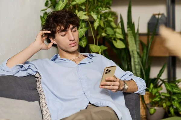 Mavi gömlekli yakışıklı genç adam koltukta dinleniyor, rahat bir yerde telefonunu karıştırıyor..