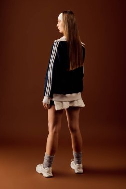 Kendine güvenen genç bir kadın atletizmini kahverengi bir zemine karşı moda spor kıyafetlerinde sergiliyor..