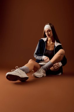 Genç bir kadın, kahverengi bir stüdyo arkaplanı içinde şık spor kıyafetleriyle rahatça poz veriyor..