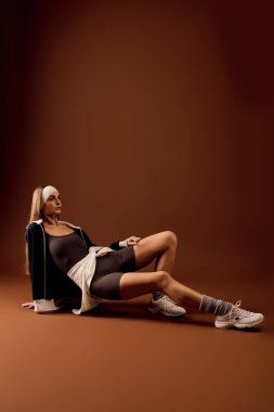 Genç bir kadın, sıcak kahverengi bir zemine karşı, kendine güveni tam bir spor elbisesi içinde poz veriyor..