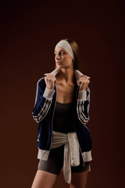 Formda genç bir kadın şık spor kıyafetleri içinde rahat kahverengi bir stüdyo arkaplanı içinde zarifçe poz veriyor..