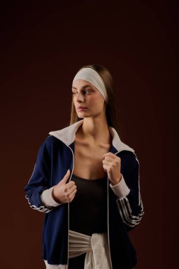 Formda genç sporcu kadın, zengin kahverengi bir arkaplana karşı şık spor kıyafetleriyle güven içinde poz veriyor..