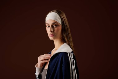 Şık spor kıyafetleri içindeki kararlı genç bir kadın kahverengi bir stüdyoda zarafetle poz veriyor..