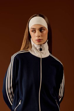 Formda genç bir kadın spor tarzını kahverengi bir stüdyo arkaplanına karşı şık bir kıyafetle sergiliyor..