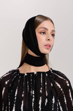 Genç bir kadın, minimalist gri bir stüdyoda zarif kıyafetler ve şık bir saç bandı sergiliyor..