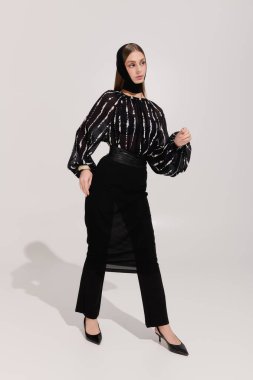 Şık giyinmiş genç bir kadın, gri bir stüdyoda güven içinde poz veriyor..