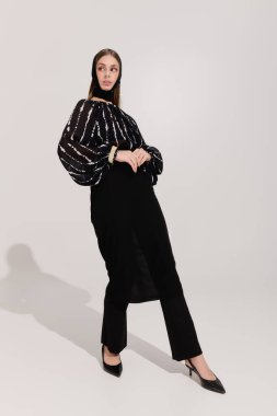 Zarif giyinmiş güzel bir genç kadın gri bir stüdyoda kendine güvenerek poz veriyor..