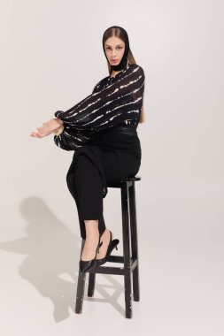 Zarif giyinmiş güzel genç bir kadın zarif bir şekilde bir taburede oturup moda anlayışını sergiliyor..