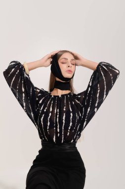 Gri bir stüdyoda zarif bir şekilde poz veren güzel bir genç kadın, şık kıyafetler ve saç bandı takıyor..