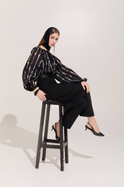 Genç bir kadın zarif bir şekilde şık giyinmiş ve gri bir arka planda şık bir saç bandıyla poz veriyor..