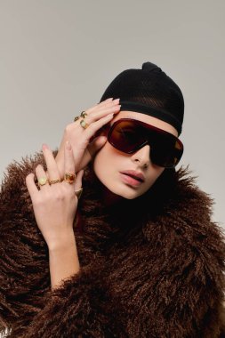 Genç bir kadın, gri bir stüdyoda poz verirken şık, sahte bir kürk mankenliği yapıyor..