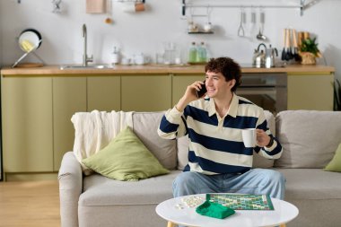 Yakışıklı bir genç adam telefonda konuşurken gülümsüyor, elinde bir fincan ve evde dinleniyor..