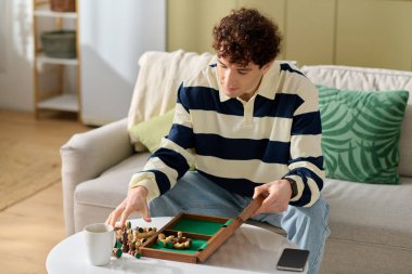Kıvırcık saçlı genç bir adam evde kahve içerken masa oyunu oynamaktan hoşlanır..
