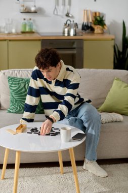 Genç bir adam, rahat bir oturma odasındaki yuvarlak bir masada siyah parçalar oyunu oynuyor..