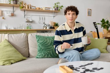 Yakışıklı genç bir adam kanepede rahatlar, elinde sıcak bir bardakla domino oynar..