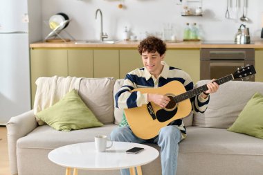 Rahat genç adam gitarını rahat bir kanepede çalar, modern bir evde kahvesini yudumlar..