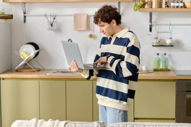 Genç adam, modern, davetkar bir mutfak alanında dururken dizüstü bilgisayarına odaklanıyor..