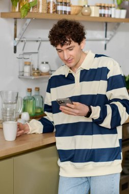 Şık bir genç adam mutfak tezgahına eğiliyor, bardağı yudumluyor ve telefonuna bakıyor..