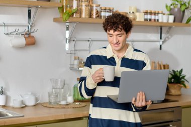Genç bir adam kahveyi yudumlarken gülümser ve laptopuyla şık, bitki dolu bir mutfakta çalışır..