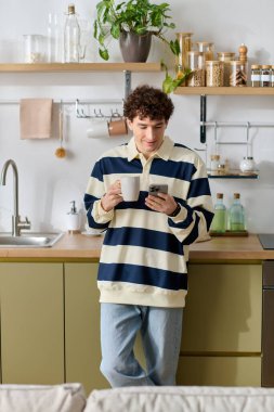 Genç bir adam sıcak kahvesini yudumluyor ve modern, davetkar bir mutfakta telefonuna bakıyor..