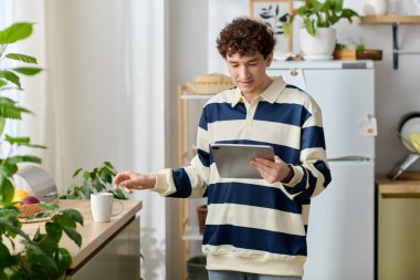 Sıcak bir mutfak ortamında, yakışıklı genç bir adam kahvesinin tadını çıkarırken tabletini kontrol eder..