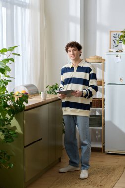 Yakışıklı genç adam, parlak bir odada, tahta bir masanın yanında kağıtları tutarken rahat duruyor..