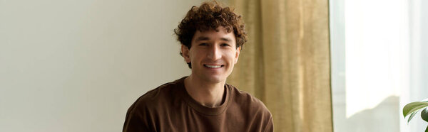 A young man with a charming smile sits relaxed at home, bathed in warm sunlight from the window.