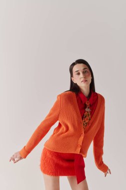 In a bold orange cardigan and red outfit, a young woman poses playfully against a simple backdrop.