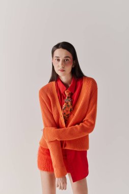 A fashionable woman poses confidently in an eye catching orange cardigan layered over a red outfit.