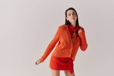 A young woman in an orange cardigan and red outfit poses creatively in a modern space.