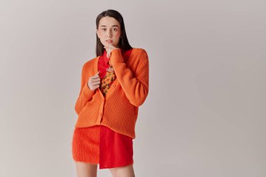 Zarif bir kadın kendine güvenerek koyu turuncu bir hırka giyiyor ve dikkat çekici kırmızı bir kıyafet giyiyor..